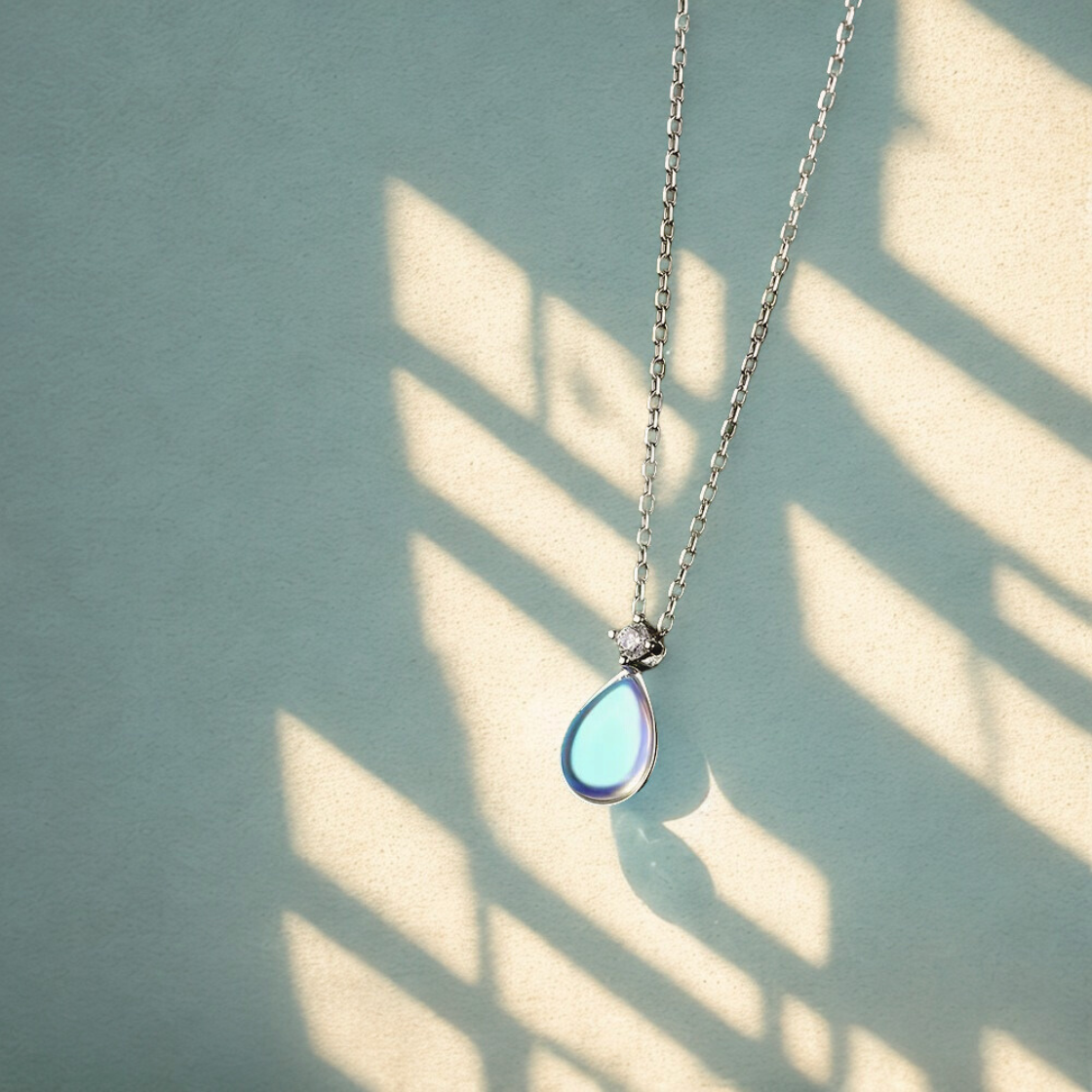 Collier argenté avec pendentif pierre de lune à reflets bleus, tenu en hauteur sur fond clair et lumineux.